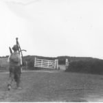 Black and white photograph showing a farm worker walking carrying his scythe and other tools