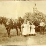 Sepia photograph showing a haymaking scene, with loaded wagon, two horses, three men, three girls and their implements