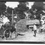 A black and white photograph of a steam engine driving a threshing machine beside straw stack