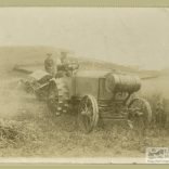 Black and white photograph of two men sat on an early tractor with farming equipment behind in a field