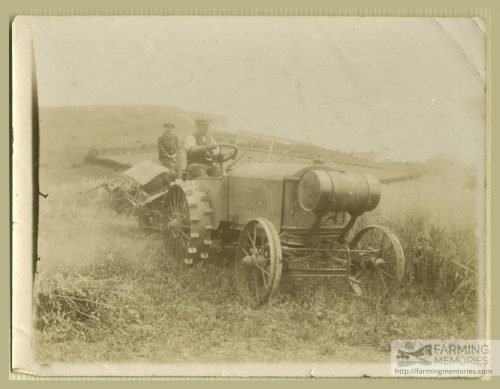 Black and white photograph of two men sat on an early tractor with farming equipment behind in a field