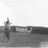 Black and white photograph showing a farm worker walking carrying his scythe and other tools