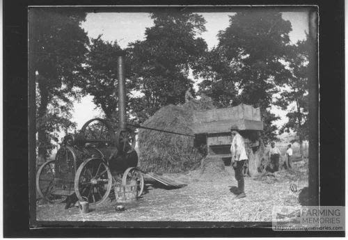 Photographic print taken from a glass plate negative in the collection of men working a threshing machine driven by a steam engine