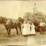 Sepia photograph showing a haymaking scene, with loaded wagon, two horses, three men, three girls and their implements