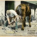 Tinted photograph shows a blacksmith shoeing a horse, with tools and a bicycle visible in the background